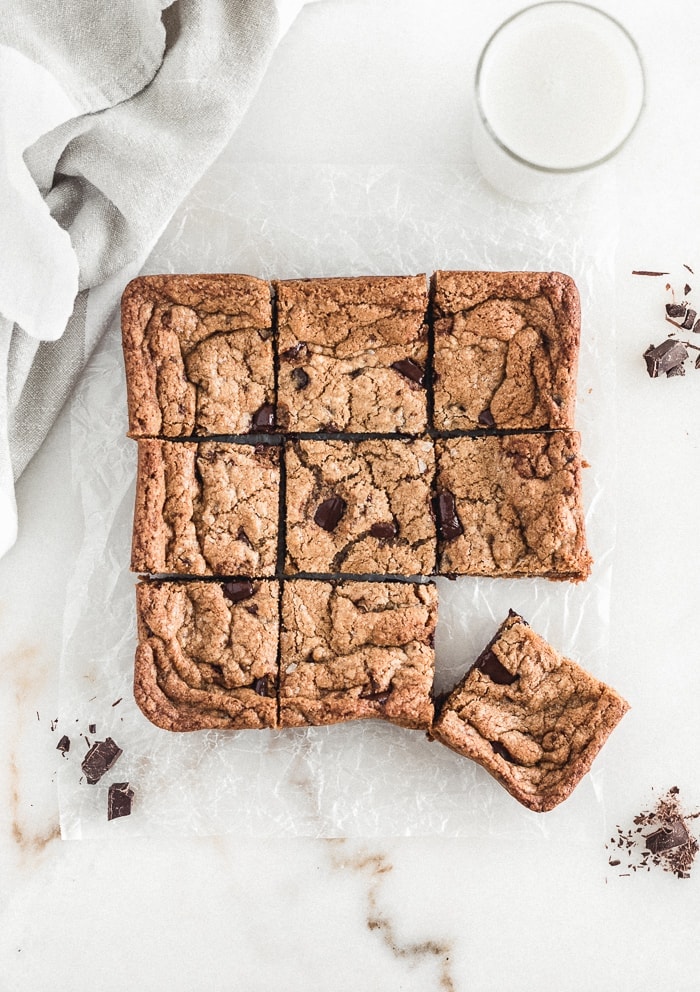 overhead view of chocolate chunk blondies cut into squares surrounded but chocolate chunks, a glass of milk, and a linen napkin.