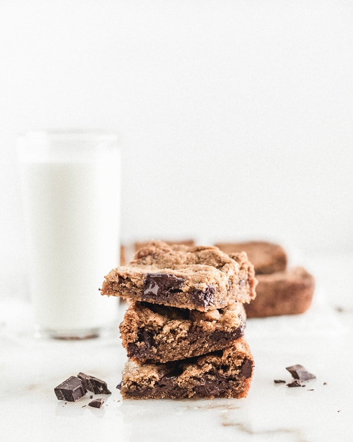 Three chocolate chunk blondies stacked on top of each other with a glass of milk and more blondies in the background.