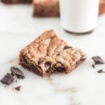 chocolate chunk blondie with a bite taken out, surrounded by chocolate chunks, with a glass of milk in the background.