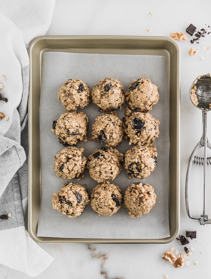 Gluten-free dark chocolate and walnut oatmeal cookie dough formed into balls on a cooking sheet.