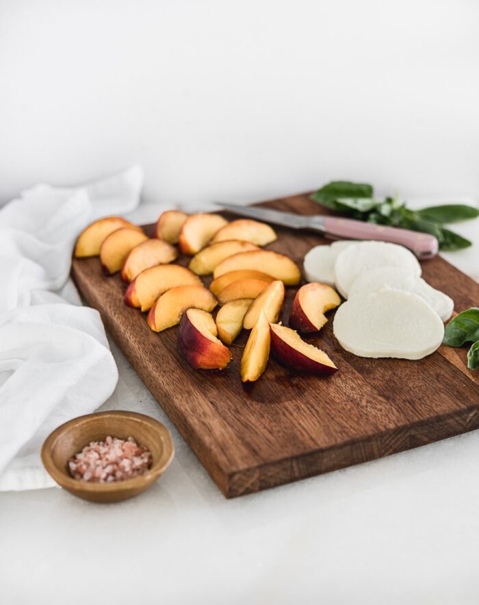 sliced peaches and sliced fresh mozzarella on a wooden cutting board with a pink knife and a bowl of pink salt next to it.