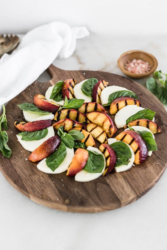 Grilled Peach Caprese salad on a round wooden cutting board with a white linen, basil and bowl of salt in the background.