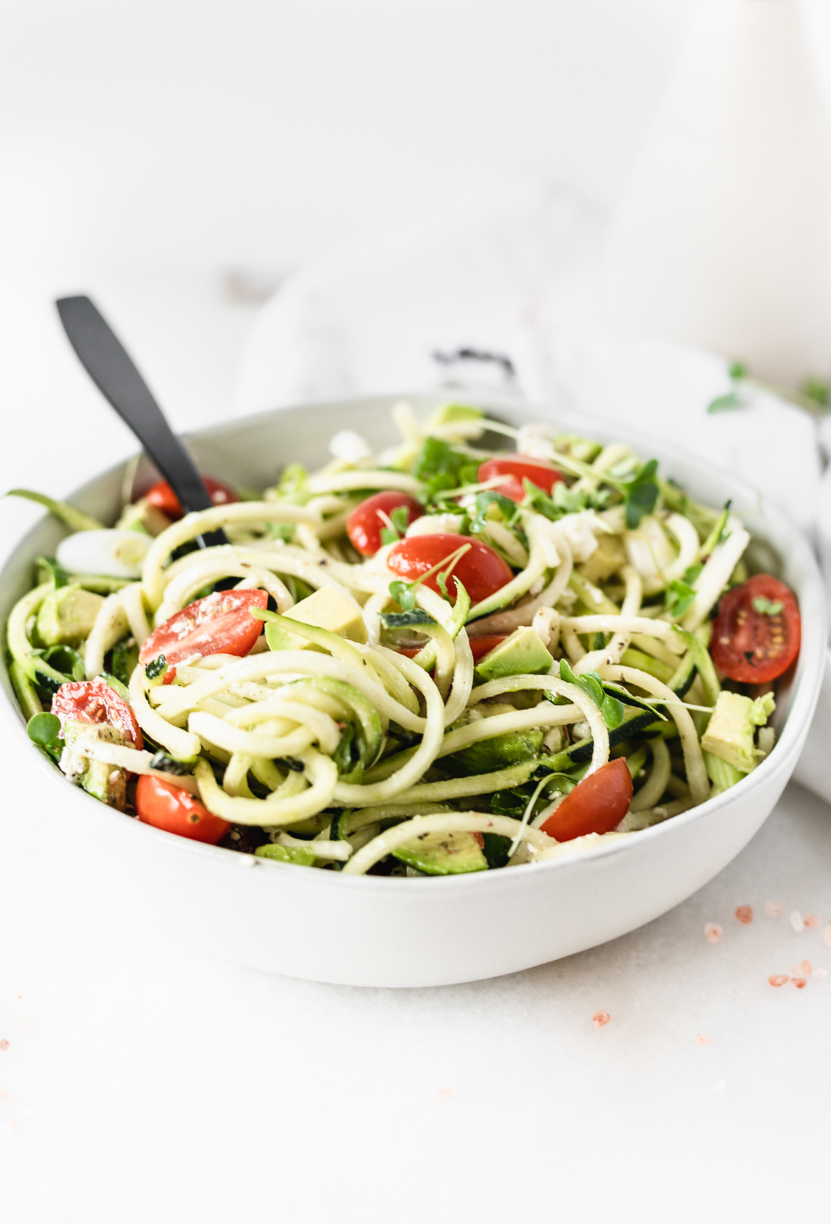 closeup of avocado tomato zucchini noodle salad in a white bowl with a black fork in it.