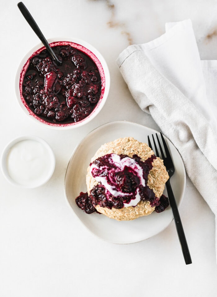 overhead view of quinoa pancakes with berry compote on a white plate, bowl of berry compote, and bowl of yogurt, and white linen.