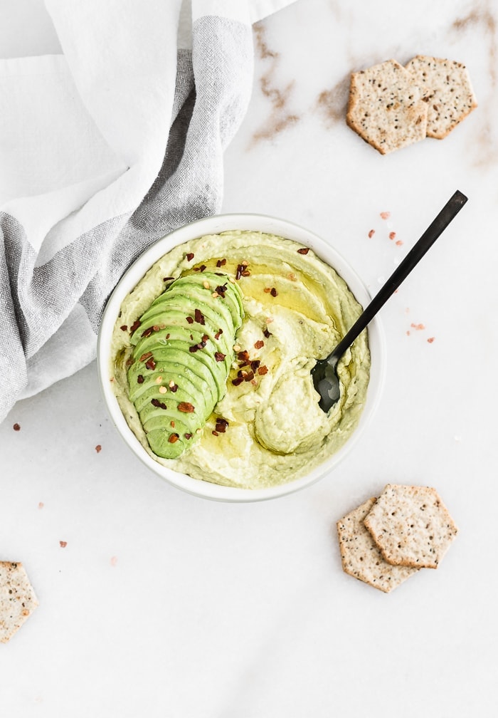 overhead view of a bowl of avocado hummus topped with sliced avocado and red pepper flakes, surrounded with crackers.