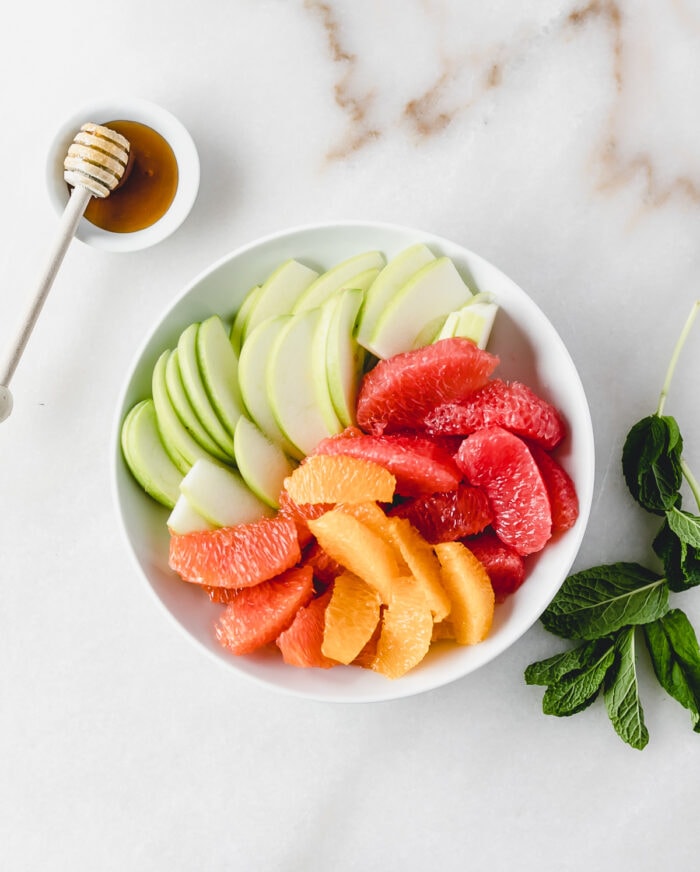 granny smith apple, navel orange, cara cara orange, and grapefruit slices places separately in a white bowl with mint and honey next to it.