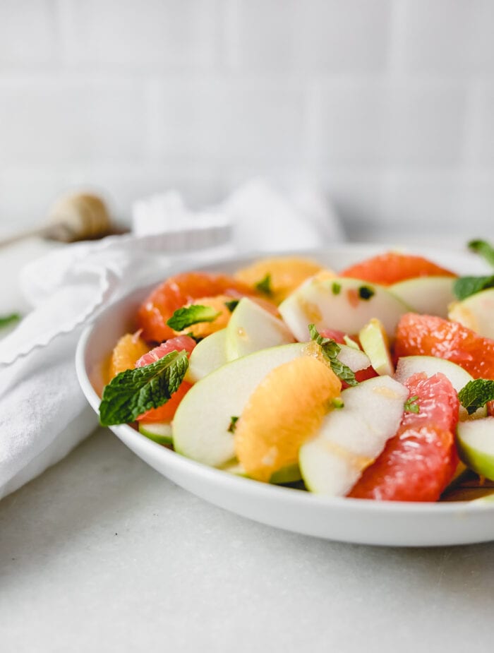 closeup of citrus apple salad in a white bowl topped with fresh mint.