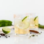hatch chile margarita in a glass with a lime slice, surrounded by lime and spices.
