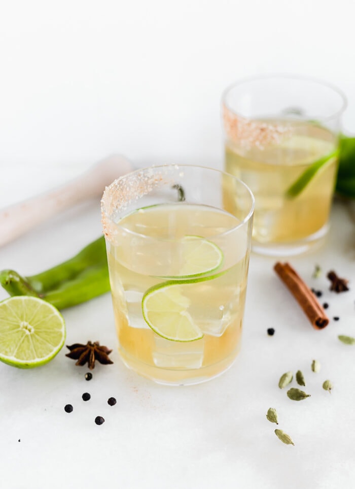 hatch chile margaritas in glasses with ice and lime slices, surrounded by hatch chiles, lime halves, and spices.