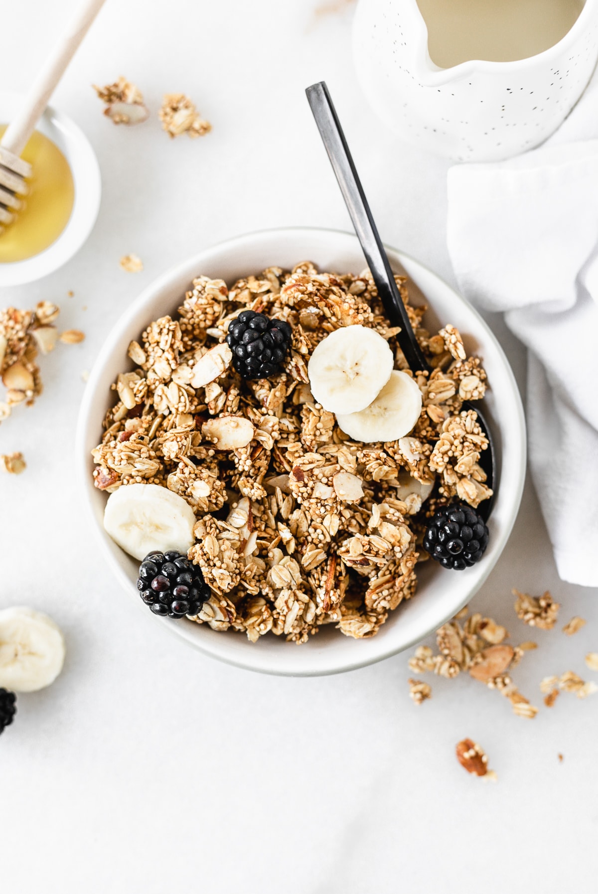 Overhead view of a bowl of honey almond quinoa granola topped with banana slices and blackberries with a black spoon in it.