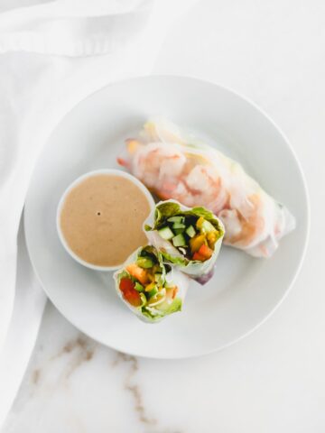 Overhead view of mango shrimp spring rolls cut in half on a white plate with Thai peanut sauce on the side.