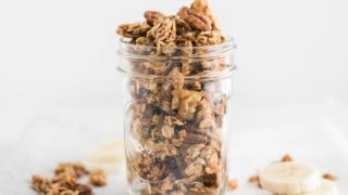 close up of healthy banana bread granola in a clear mason jar on top of a napkin with granola and banana slices on the side.