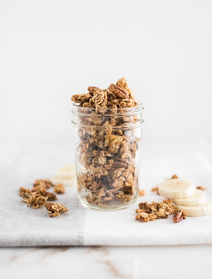 close up of healthy banana bread granola in a clear mason jar on top of a napkin with granola and banana slices on the side.