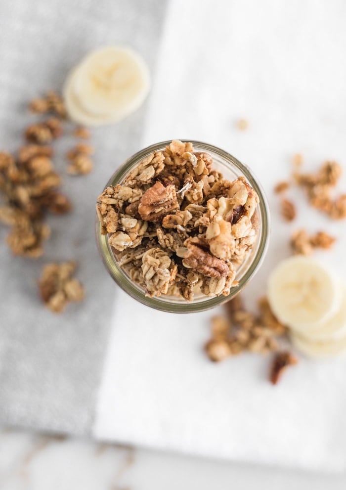 top view of healthy banana bread granola in a clear mason jar on top of a napkin with granola and banana slices on the side.