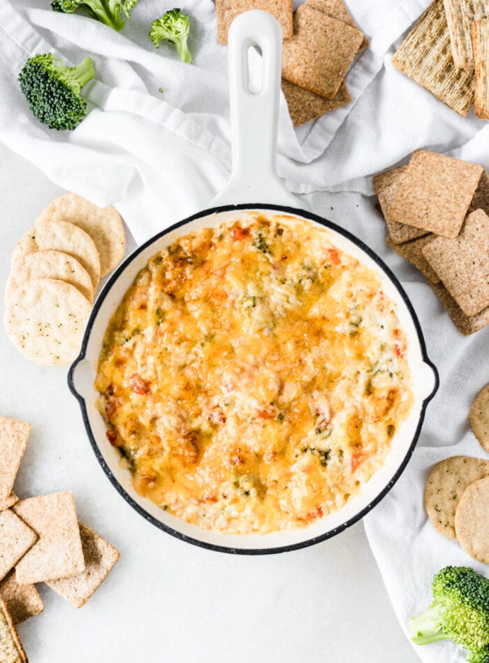 overhead view of cheesy healthy hot broccoli dip in a white skillet surrounded by crackers and broccoli florets.