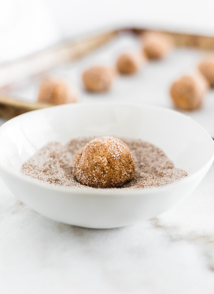ball of pumpkin snickerdoodle dough being rolled in cinnamon sugar in a white bowl with a tray of cookie dough behind it.