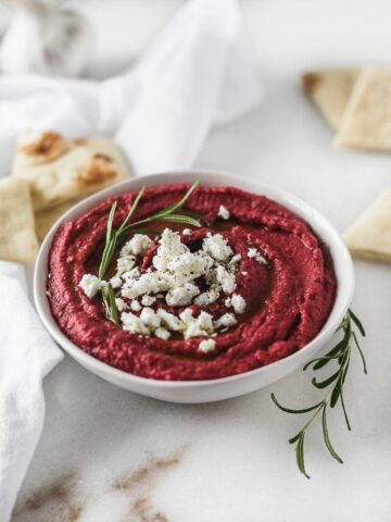 Roasted beet hummus in a white bowl garnished with goat cheese and fresh rosemary.