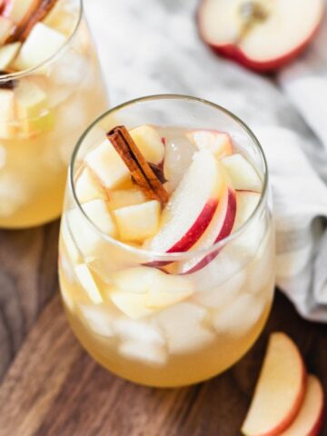 apple cider sangria in a glass with apple slices and a cinnamon stick on a wooden board with a napkin and cut apple.