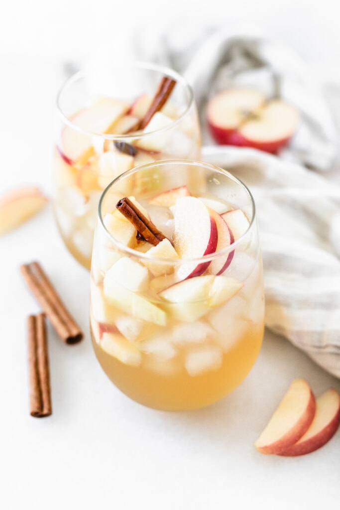 two glasses of apple cider sangria garnished with cinnamon sticks and apple slices surrounded by a grey striped linen, cinnamon sticks and apples.