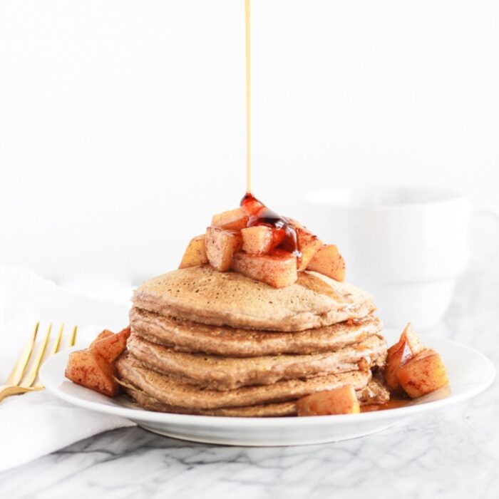 healthy apple cinnamon pancakes with maple syrup being poured on top.