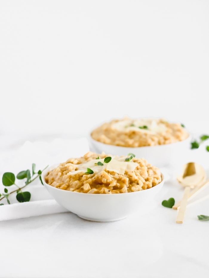 creamy pumpkin risotto in a white bowl topped with parmesan and herbs in front of another bowl of risotto.