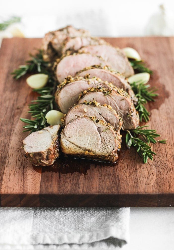 rosemary garlic pork tenderloin sliced on a wooden cutting board with garlic and herbs.