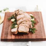 sliced pork tenderloin on a wooden cutting board surrounded by rosemary sprigs and garlic cloves.