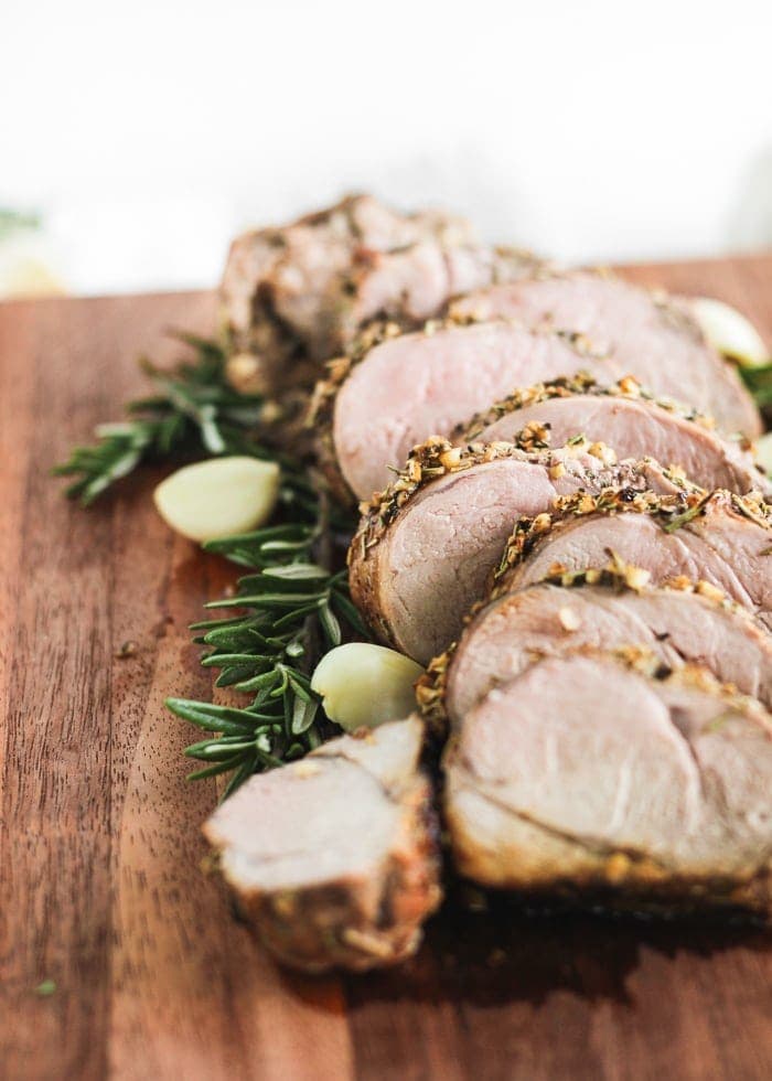 closeup view of rosemary garlic pork tenderloin sliced on a wooden cutting board with garlic and herbs.