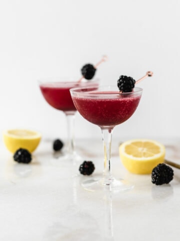two blackberry gin and jam cocktails in coupe classes surrounded by lemons and blackberries.