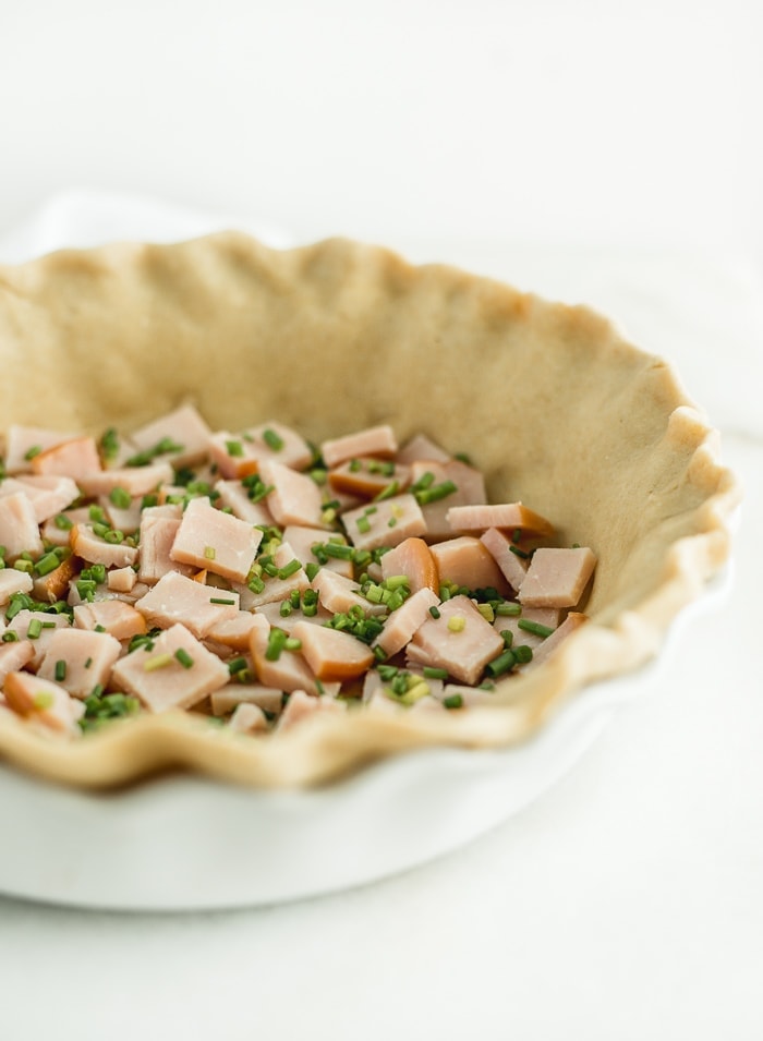 chopped canadian bacon and chives in an uncooked pie crust in a white pie dish.