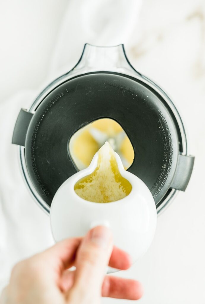 hand pouring melted butter from a white dish into a blender to make hollandaise sauce.