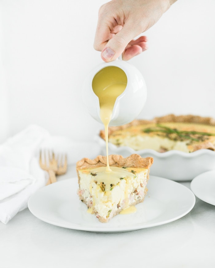 hand pouring hollandaise sauce over a slice of eggs benedict quiche on a white plate.