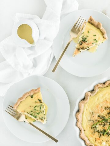 Overhead view of two slices of Eggs Benedict Quiche on white plates with gold forks, a pouring dish of hollandaise sauce, and a pie dish of quiche.