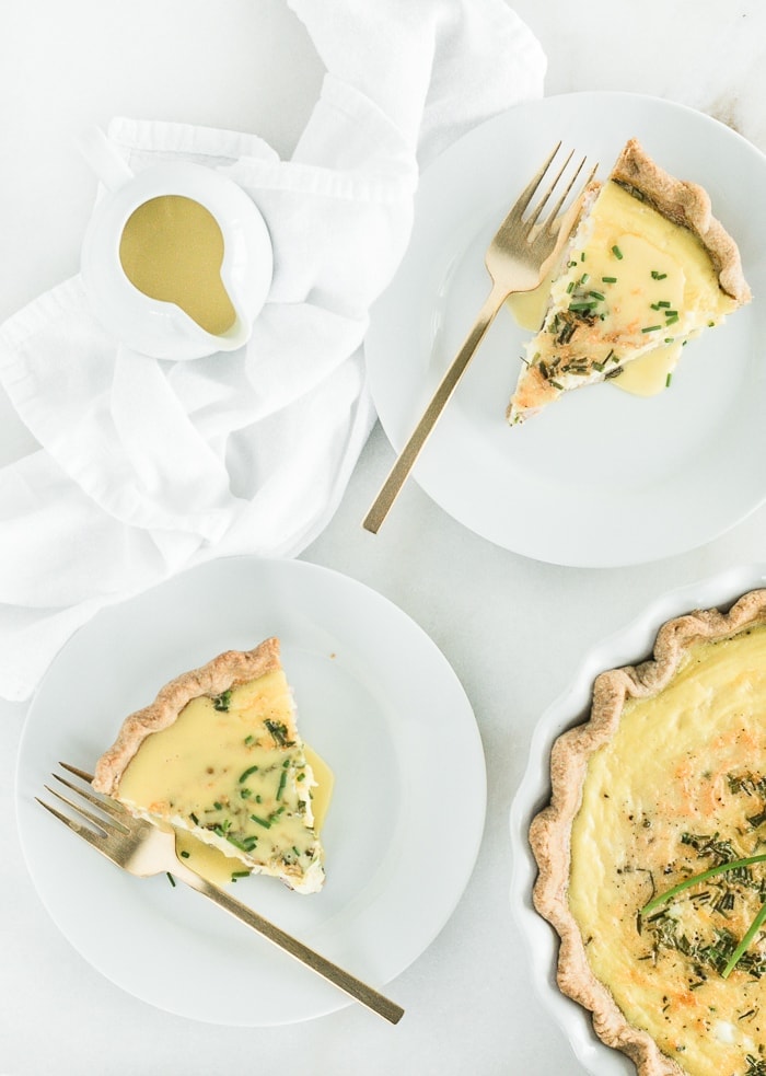 Overhead view of two slices of Eggs Benedict Quiche on white plates with gold forks, a pouring dish of hollandaise sauce, and a pie dish of quiche.