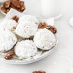 pecan snowball cookies on a plate with pecans with milk and a bowl of pecans in the background.