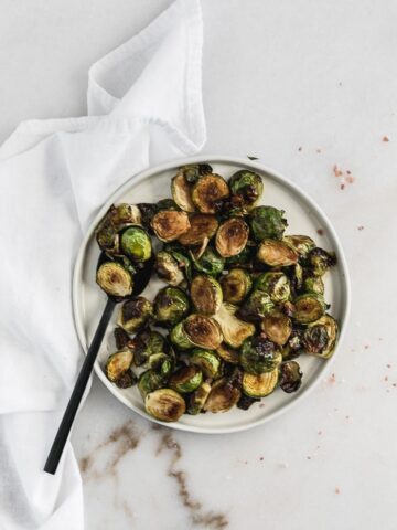overhead shot of simple balsamic roasted brussels sprouts on a white plate with a black spoon surrounded by pink salt and a white napkin.