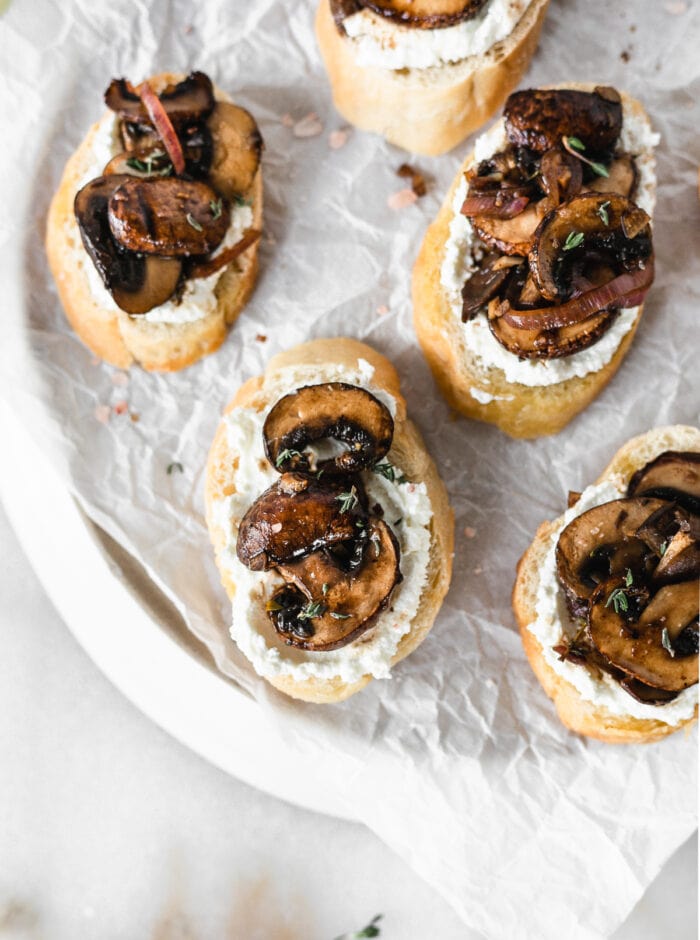 overhead view of mushroom ricotta crostini on a parchment lined plate on a white marble background.