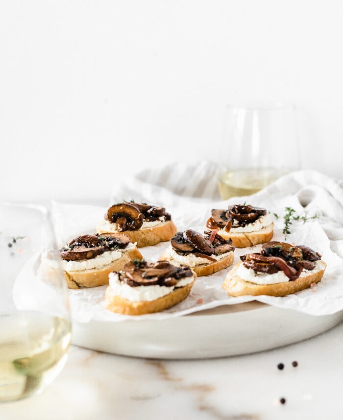 mushroom ricotta crostini on a plate lines with parchment surrounded by white wine glasses.