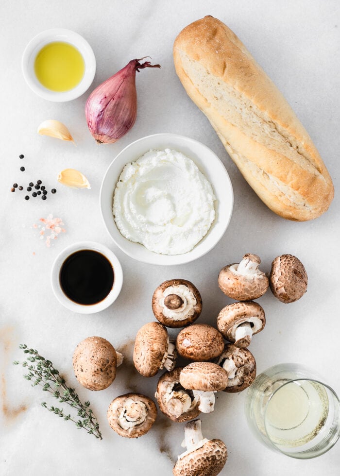 overhead view of the ingredients needed to make mushroom ricotta crostini on a white marble background.