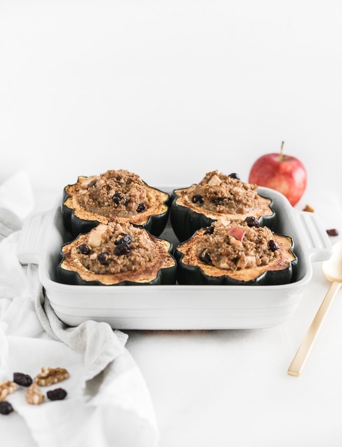 four apple quinoa stuffed acorn squash in a white square baking dish with a gold spoon beside it.