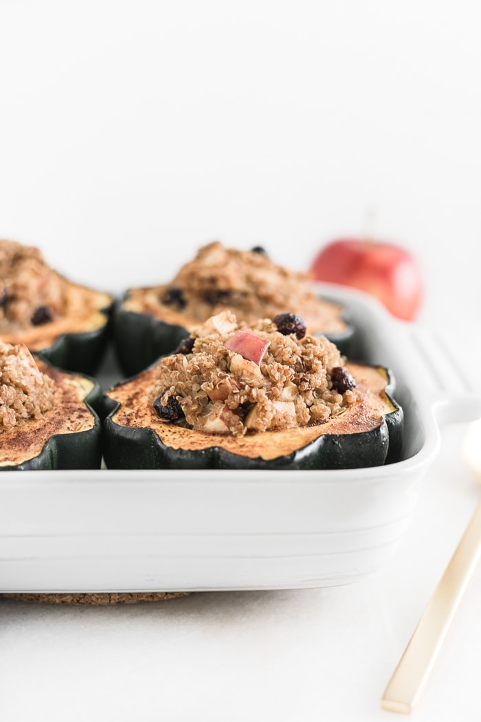 closeup of a roasted acorn squash half stuffed with quinoa, apples and cranberries in a white baking dish.