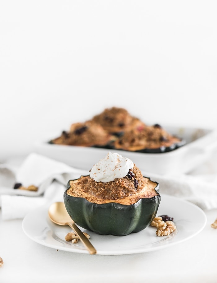 Roasted acorn squash half stuffed with quinoa filling and topped with yogurt on a white plate with a gold spoon beside it. 