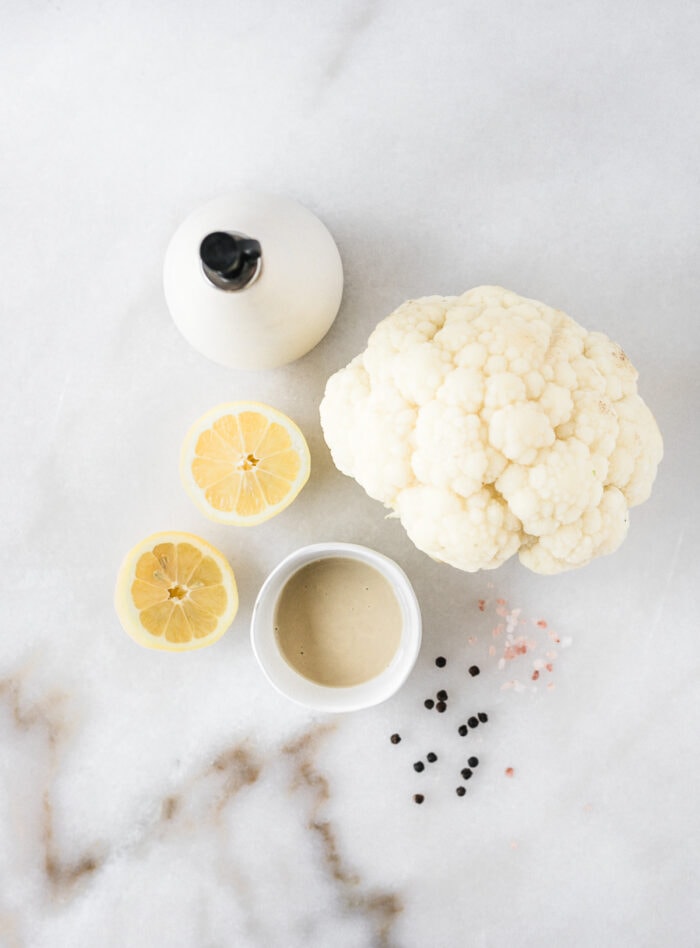 overhead view of a head of cauliflower, bottle of olive oil, bowl of tahini, pink salt and peppercorns.