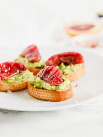 Close up view of four blood orange avocado crostini on a white serving plate.