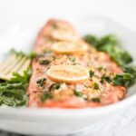 close up of garlic cilantro salmon topped with herbs and lemon slices on a white plate.
