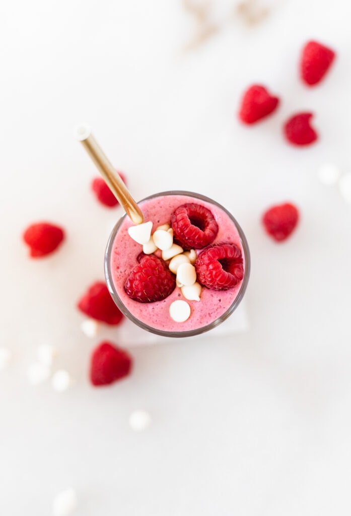 overhead view of a healthy white chocolate raspberry Valentine's smoothie with white chocolate chips and raspberries on top and around the glass.
