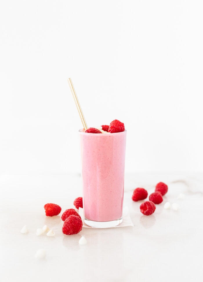 healthy white chocolate raspberry smoothie in a glass surrounded by raspberries and white chocolate chips.