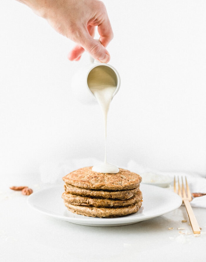 healthier cream cheese frosting being poured on a stack of carrot cake pancakes.