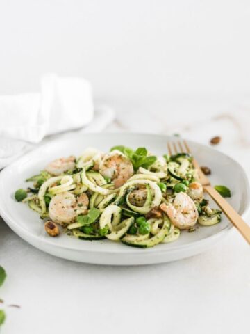 zucchini noodles with shrimp, peas and mint pesto on a white plate with a gold fork.