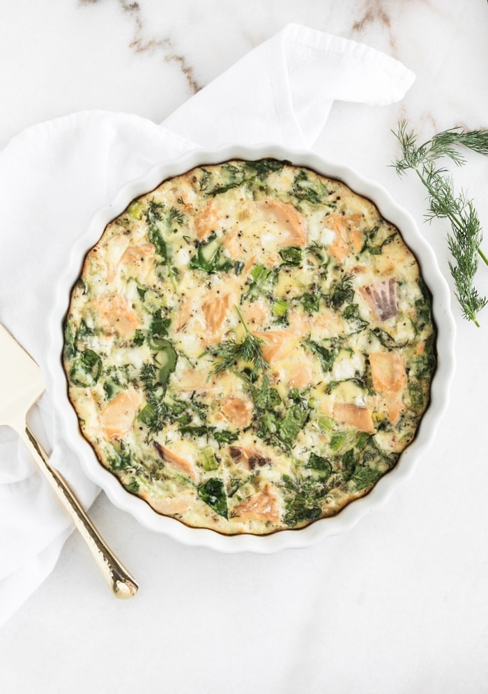 overhead view of a smoked salmon and kale frittata in a white frittata dish under a white napkin.
